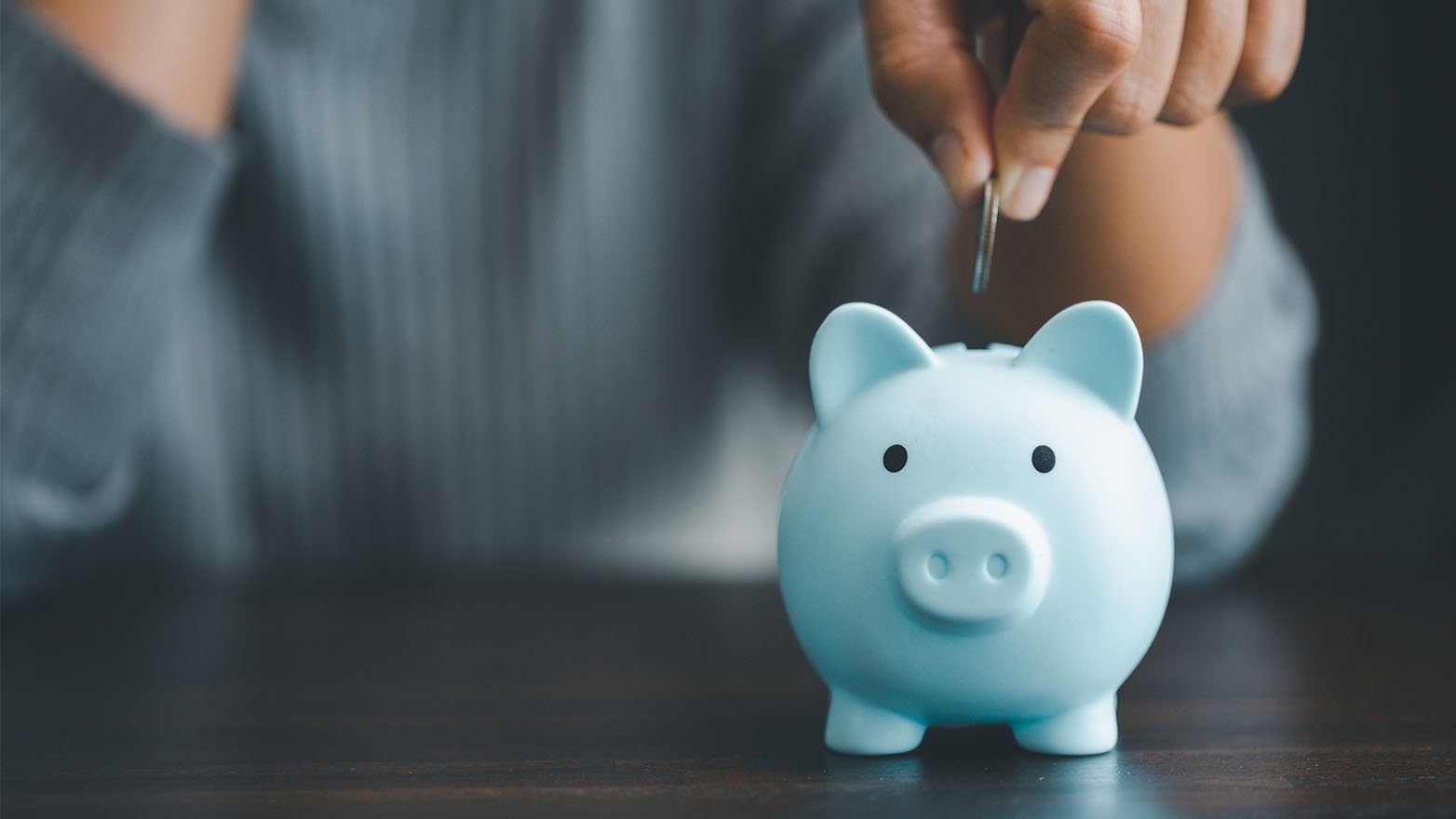 A hand releases a coin into a light blue piggy bank with a slot on top.