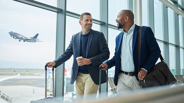 Two businessmen in suits, carrying luggage, walk and talk in an airport terminal as a plane takes off in the background, visible through a large window.