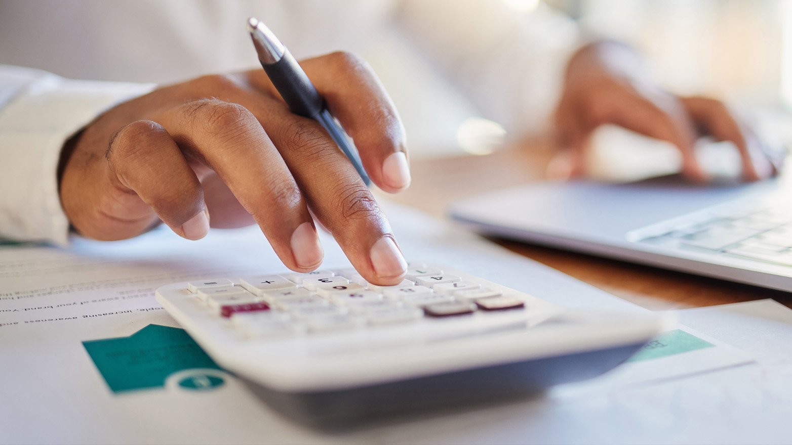 A person's hand holds a calculator and a pen, with a laptop visible in the blurred background, suggesting work or study in progress.