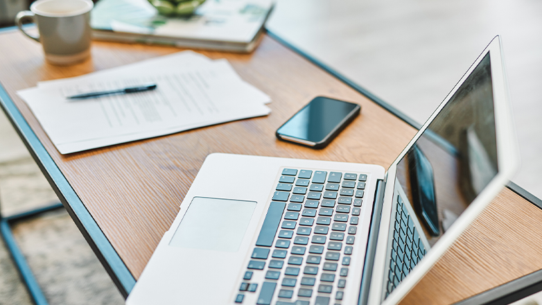 A laptop computer sits open on a wood-grain desk. The laptop screen reflects the ceiling and other objects in the room. A smartphone and some papers with a pen resting on them are next to the computer. A mug sits just out of focus at the top of the frame.