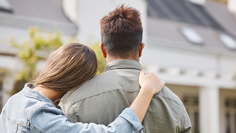 A woman rests her head on her partner's shoulder as they look at a large house.