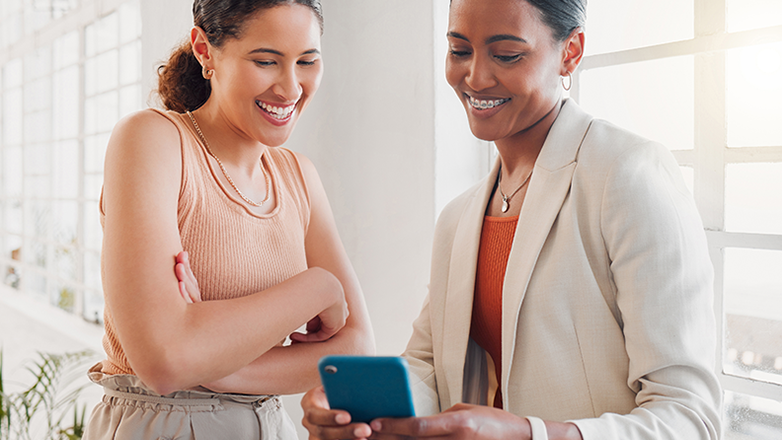 Two young women are looking at something on a smartphone together. They are both smiling and appear to be enjoying themselves.