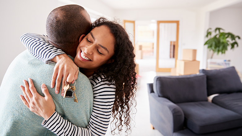 A young couple is hugging each other. The woman is holding a set of keys in her hand. They are standing in a living room that is mostly empty, except for a couch and a few boxes.