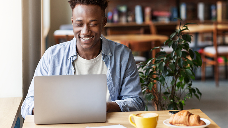A young African-American man is sitting at a table in a coffee shop, smiling and looking at his laptop. He has a cup of coffee and a croissant on the table.