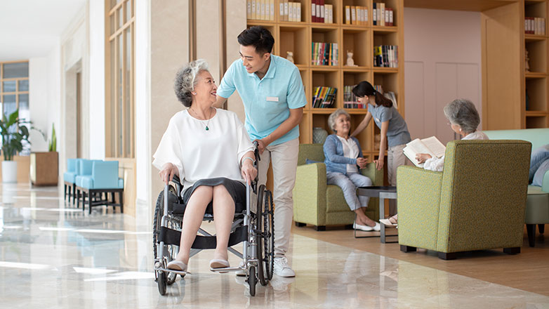Woman in wheelchair is being helped by a male assistant in the lobby of a nursing home.