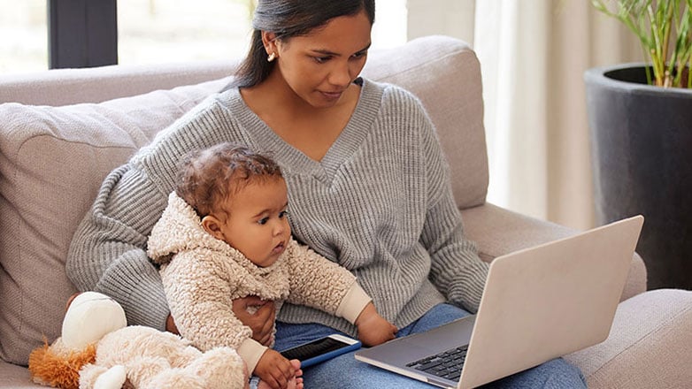 Mom cuddles with baby on a couch while working on a laptop.