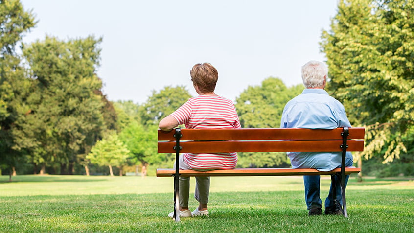 senior couple sitting on opposite sides of a bench in a park, ignoring each other.