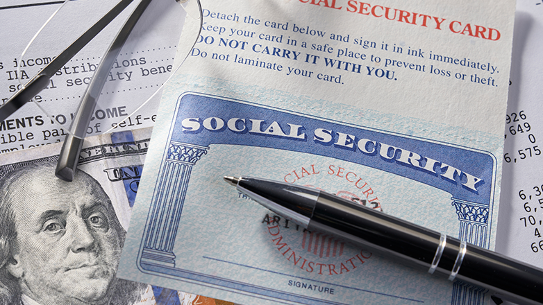 A Social Security card lays on top of various financial documents and a one hundred dollar bill. Eyeglasses and a pen rest on top of the card.