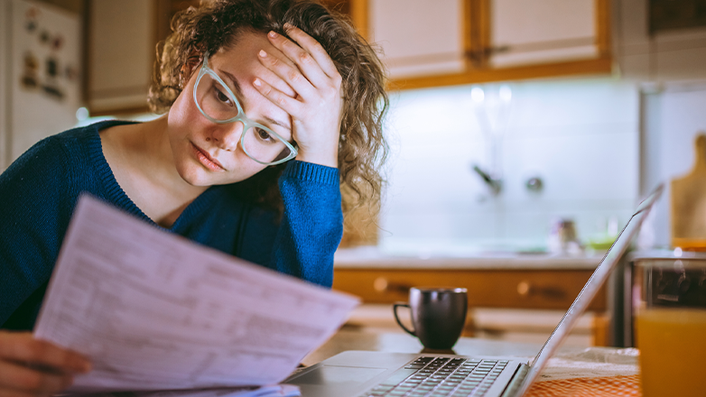 A young woman with curly hair and glasses sits at a kitchen counter, looking stressed as she reads paperwork. She rests her hand on her forehead, with a laptop open beside her.