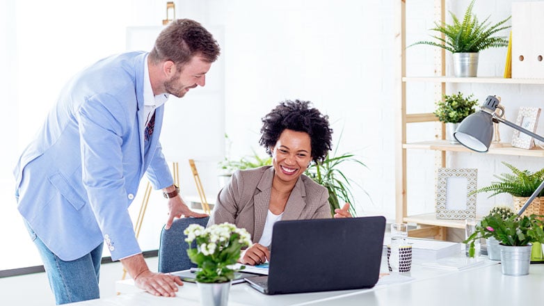 woman sitting at desk smiling looking at laptop with man standing to her side