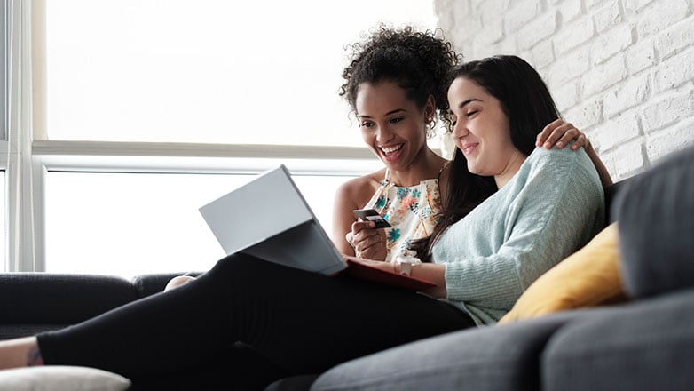 Two young females sitting on a couch making an online purchase.