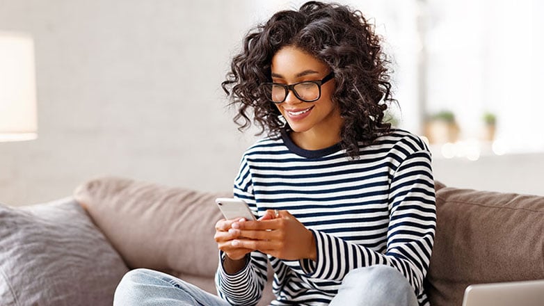 Young woman uses online banking on her phone while relaxing on her sofa.