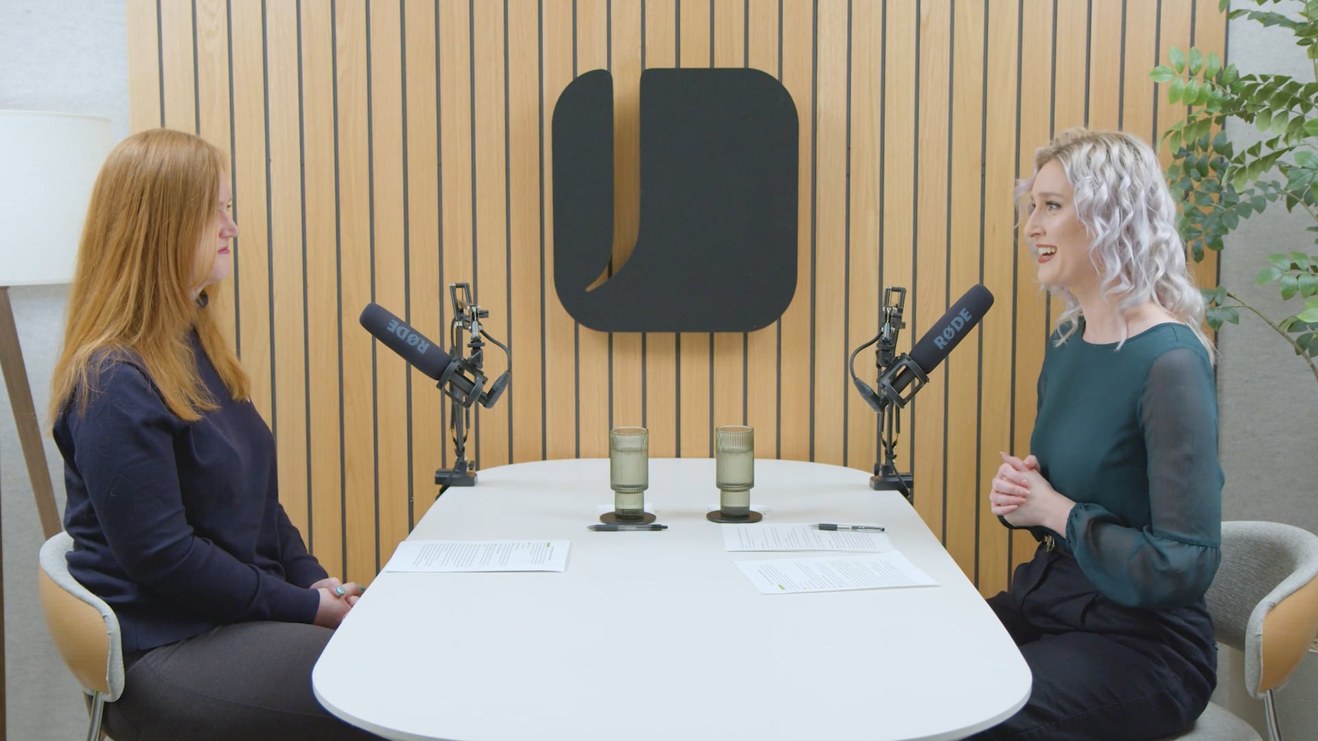 Podcast-style interview setup with two people seated across a table, speaking into microphones with notes in front of them, discussing Social Security and retirement planning topics in a modern studio setting.