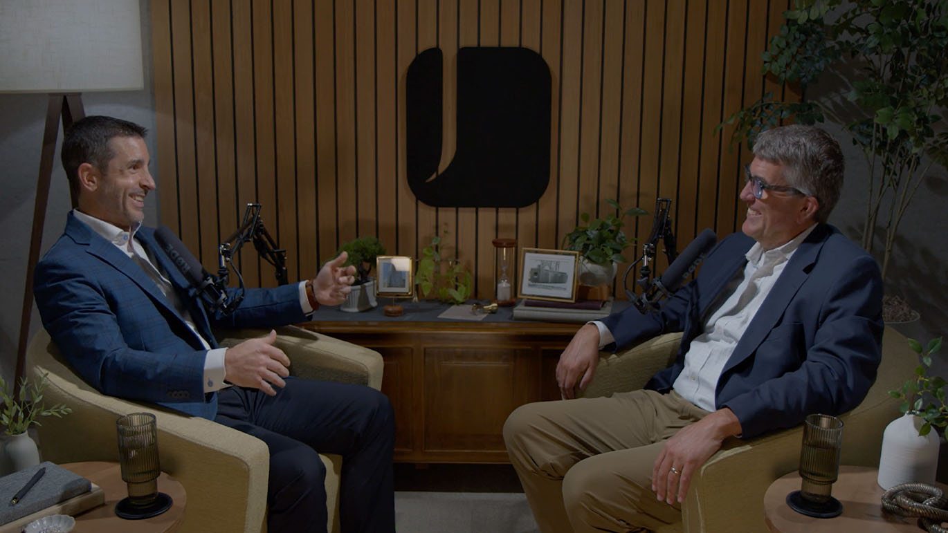 Two podcast hosts sit across from each other in armchairs, speaking into microphones in a studio with wood-paneled walls, plants, and a central table.