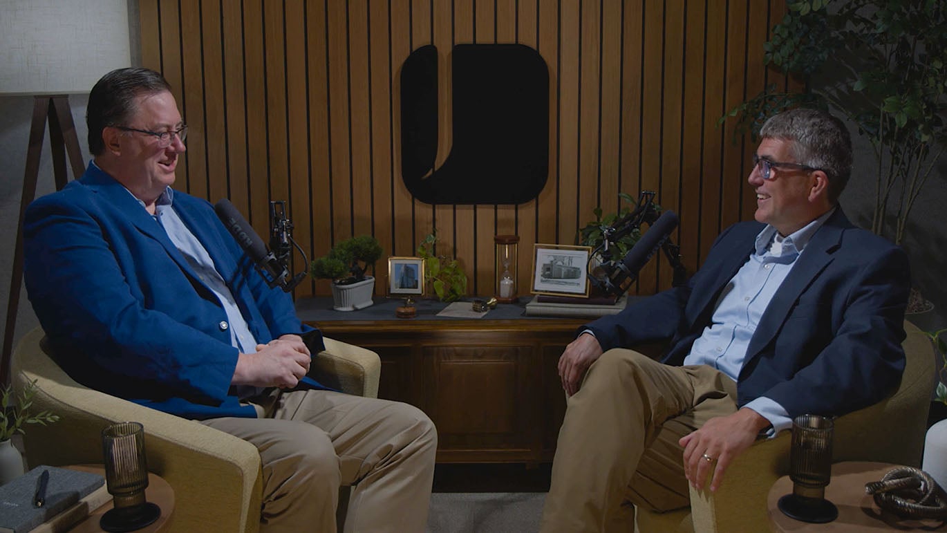 Wide shot of a podcast recording studio featuring two speakers seated across from one another, speaking into microphones with wood paneling, plants, and framed artwork in the background.