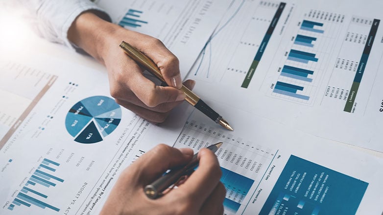 close up on two hands with pens pointing at multiple papers with graphs