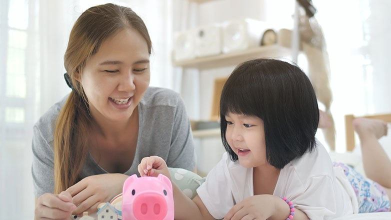 Mom and daughter laying on bed putting money in the child's piggy bank.