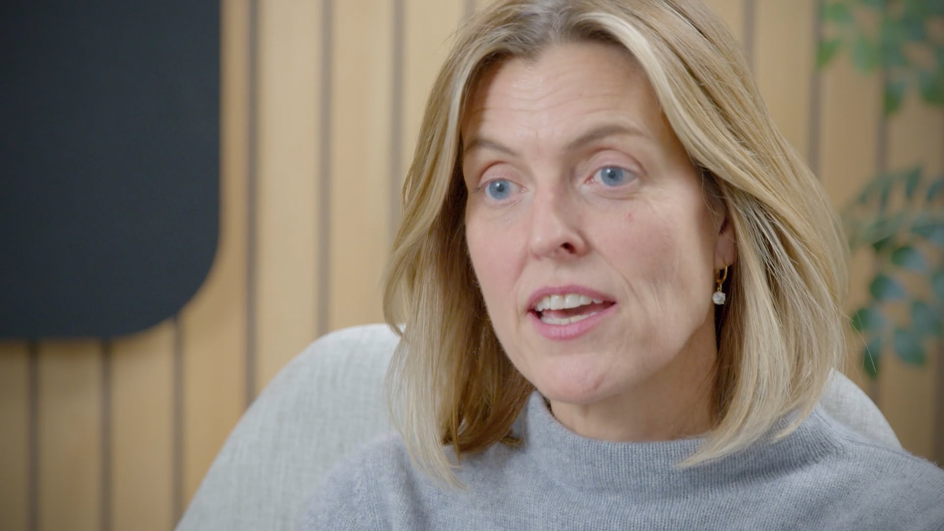 Close-up of a person with shoulder-length light-colored hair wearing a light gray sweater and a small drop earring, seated indoors with a softly blurred background of wood paneling and greenery.