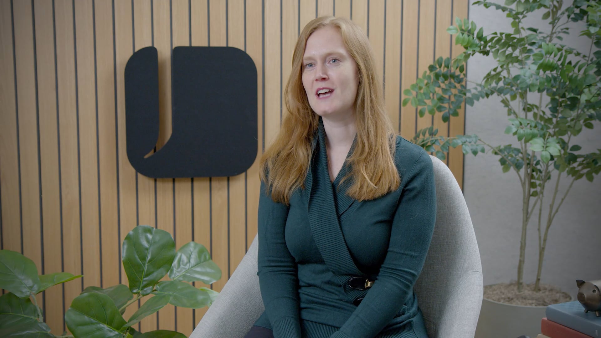 Person seated in a light gray chair in an office setting with wood slat walls, indoor plants, and a black company logo displayed behind them. The person is wearing a dark green sweater and appears to be speaking during an interview or recorded conversation.