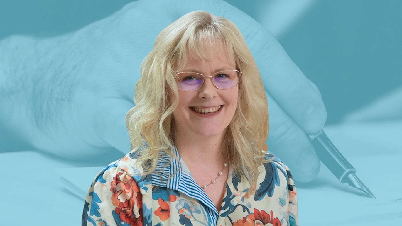 Professional headshot of a woman with shoulder-length blonde hair and glasses, wearing a floral blouse and pearl necklace. The portrait is set against a blue-toned background featuring a close-up image of a hand writing on paper with a fountain pen.
