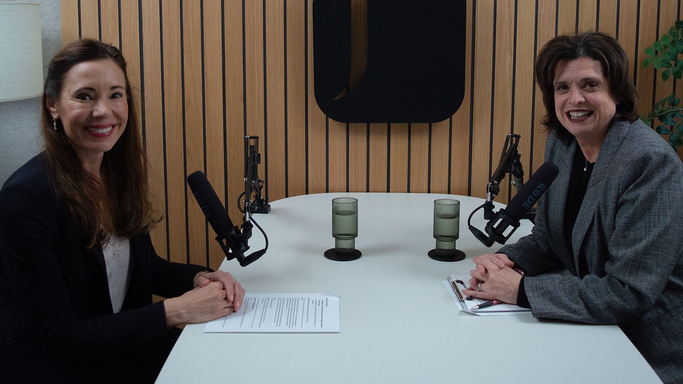 Two women smile at each other across a table in a recording studio, with microphones and glasses of water between them.