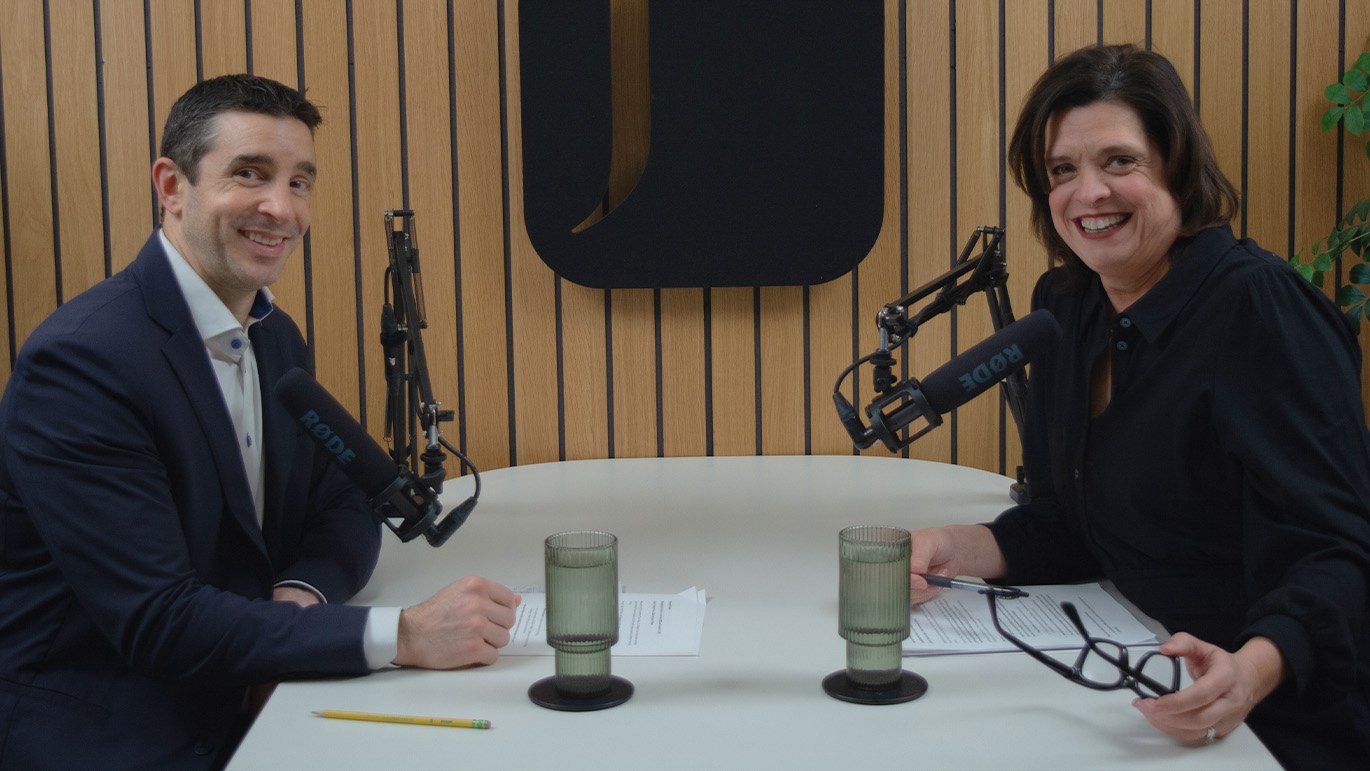 A smiling man and woman host a podcast or broadcast, seated at a table with professional microphones.