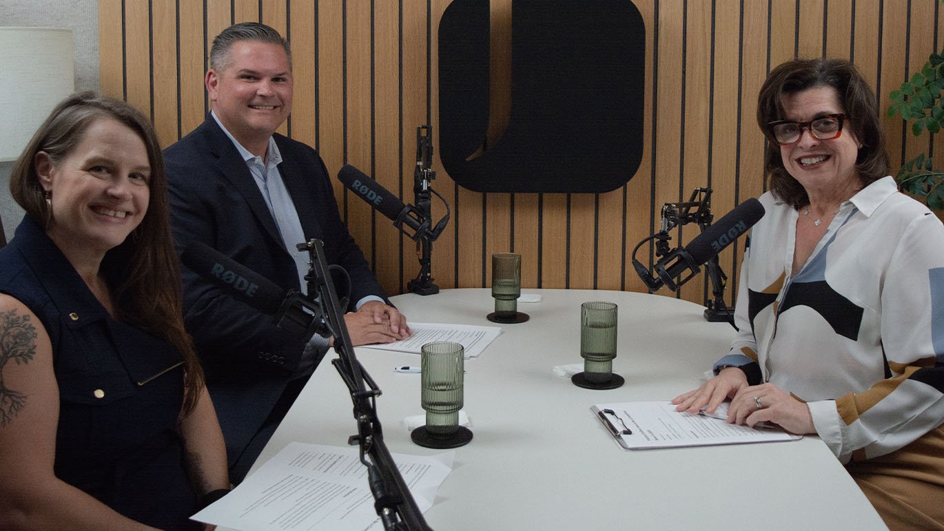 Three people—two women and one man—sit at a white table with RØDE microphones in a studio with vertical wooden slat walls. The woman on the left wears a dark sleeveless top and has an arm tattoo. The man in the center wears a dark suit. The woman on the right wears glasses and a white blouse with geometric patterns. Papers and ribbed glass tumblers are on the table.