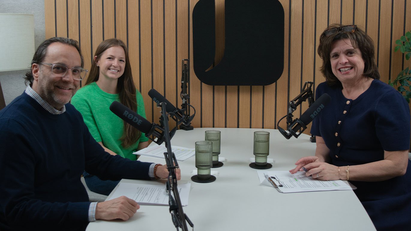 Three adults seated around a white table in a recording studio with microphones on adjustable arms. Papers and glasses of water are on the table. Vertical wood paneling and a black wall panel are visible in the background.