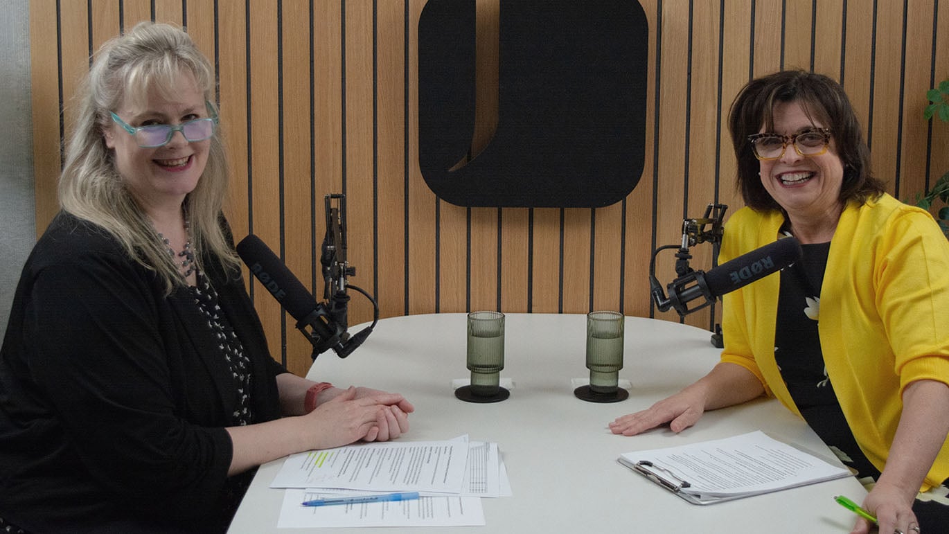 Two people sit across a small table in a recording space, speaking into microphones. Printed notes and two glasses of water are on the table, with a wood-paneled background.
