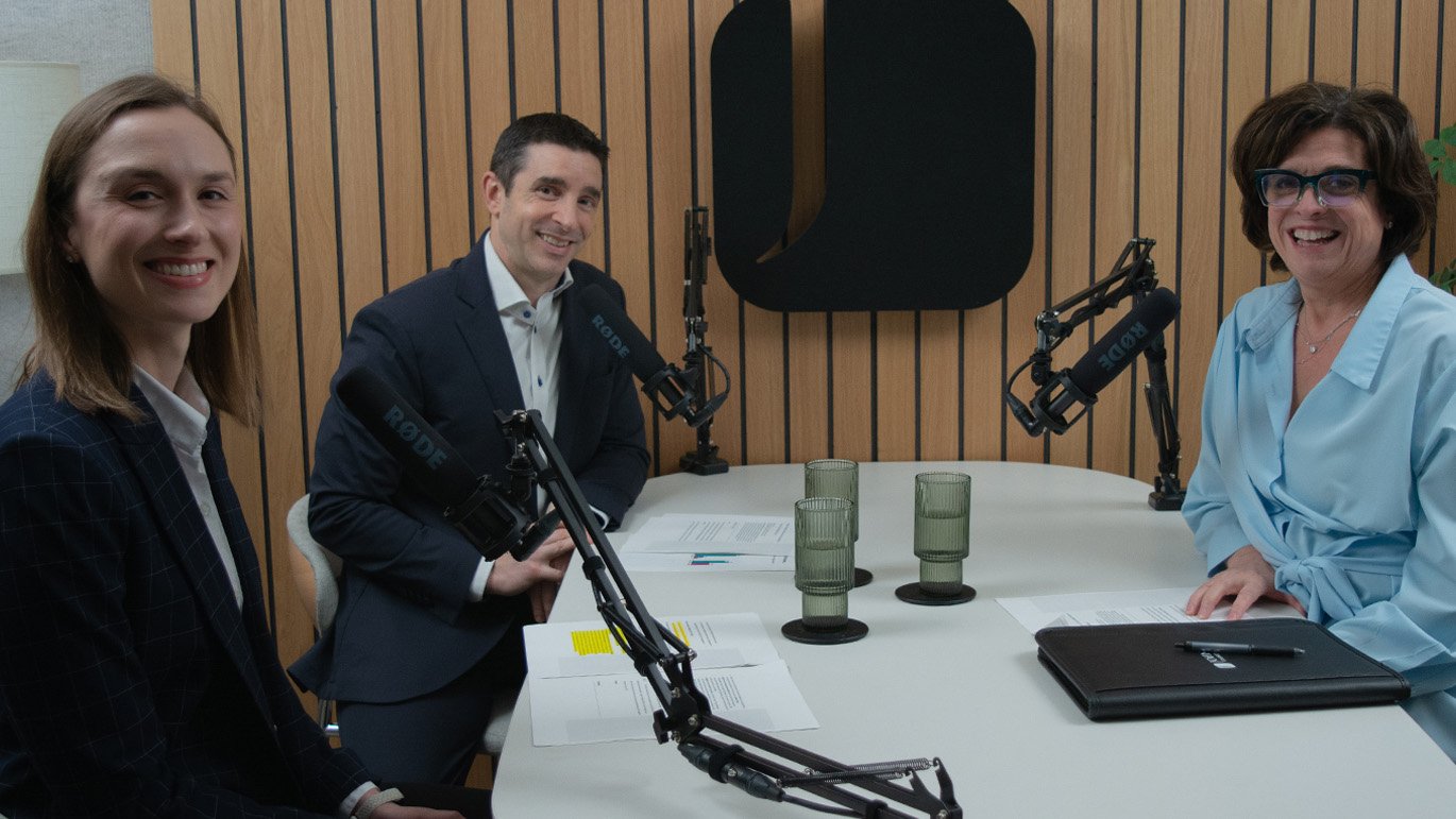 Two women and a man smile while seated at a white table in a podcast studio, with microphones and papers in front of them.