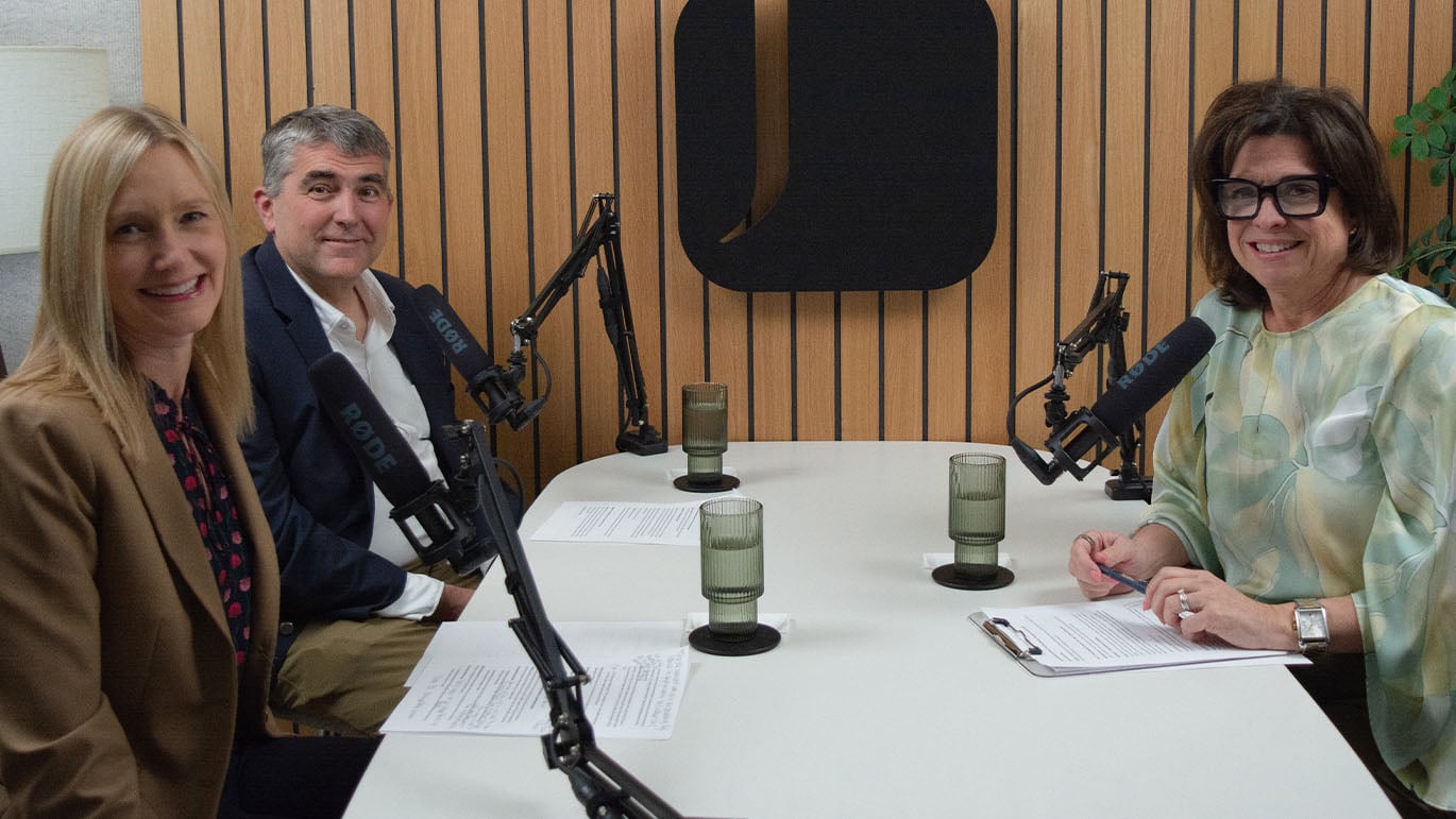 Three people seated at a podcast table with microphones and notes, recording a discussion about the great wealth transfer in a modern studio setting.