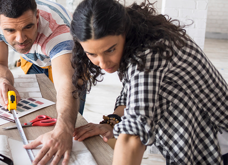 A man and woman work together on a home renovation, using a measuring tape and color swatches to plan a design. They are leaning over a table with wallpaper samples.
