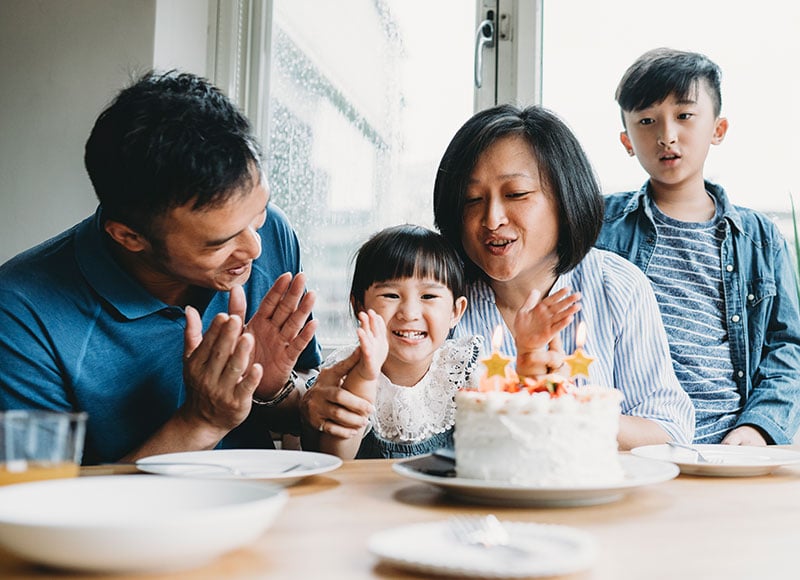 Parents sing happy birthday to their little girl as she happily claps and looks at her birthday cake.