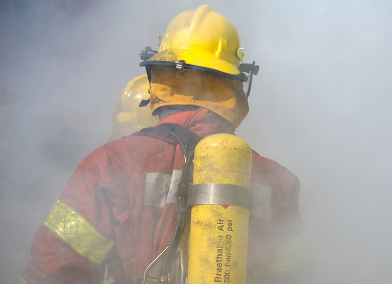 A firefighter's back is visible, wearing a yellow helmet, red turnout gear with yellow reflective strips, and a yellow air tank strapped to their back. The scene is filled with thick smoke, suggesting an active fire. Another firefighter's helmet is partially visible in the background.