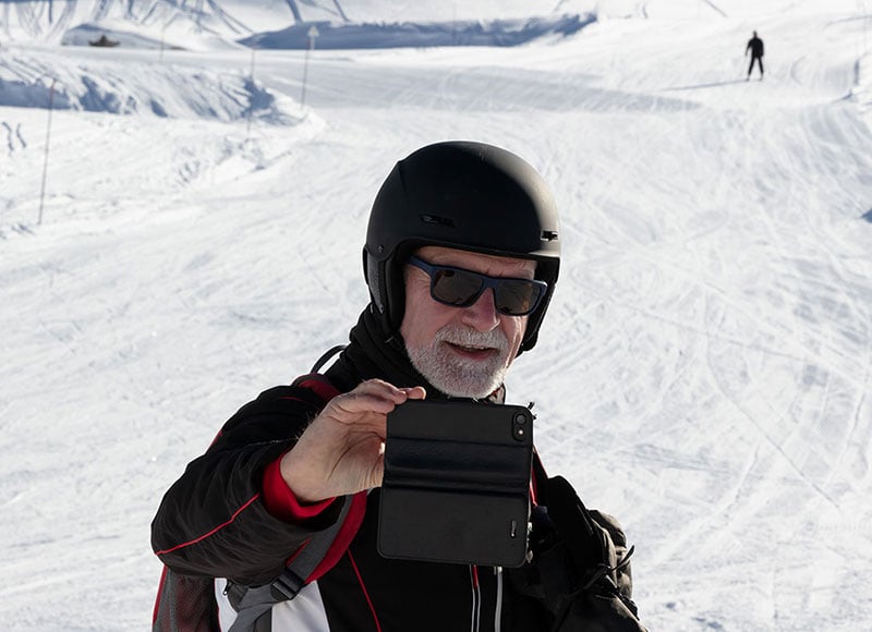 Retired man takes a selfie in front a ski slope while wearing a snowsuit.