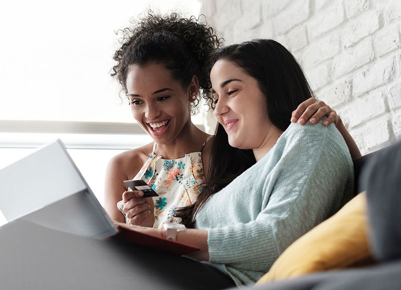 Two women sitting on a couch together and looking on a laptop.