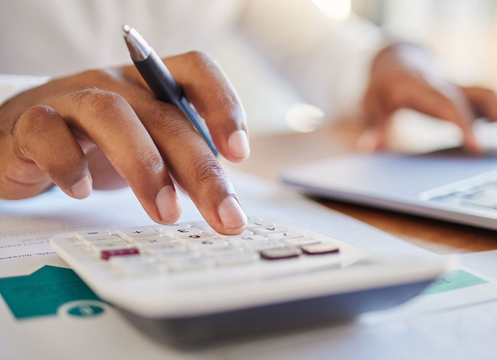 A person's hand holds a calculator and a pen, with a laptop visible in the blurred background, suggesting work or study in progress.