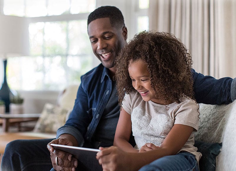 Dad and daughter sit on couch together and watch a tablet.