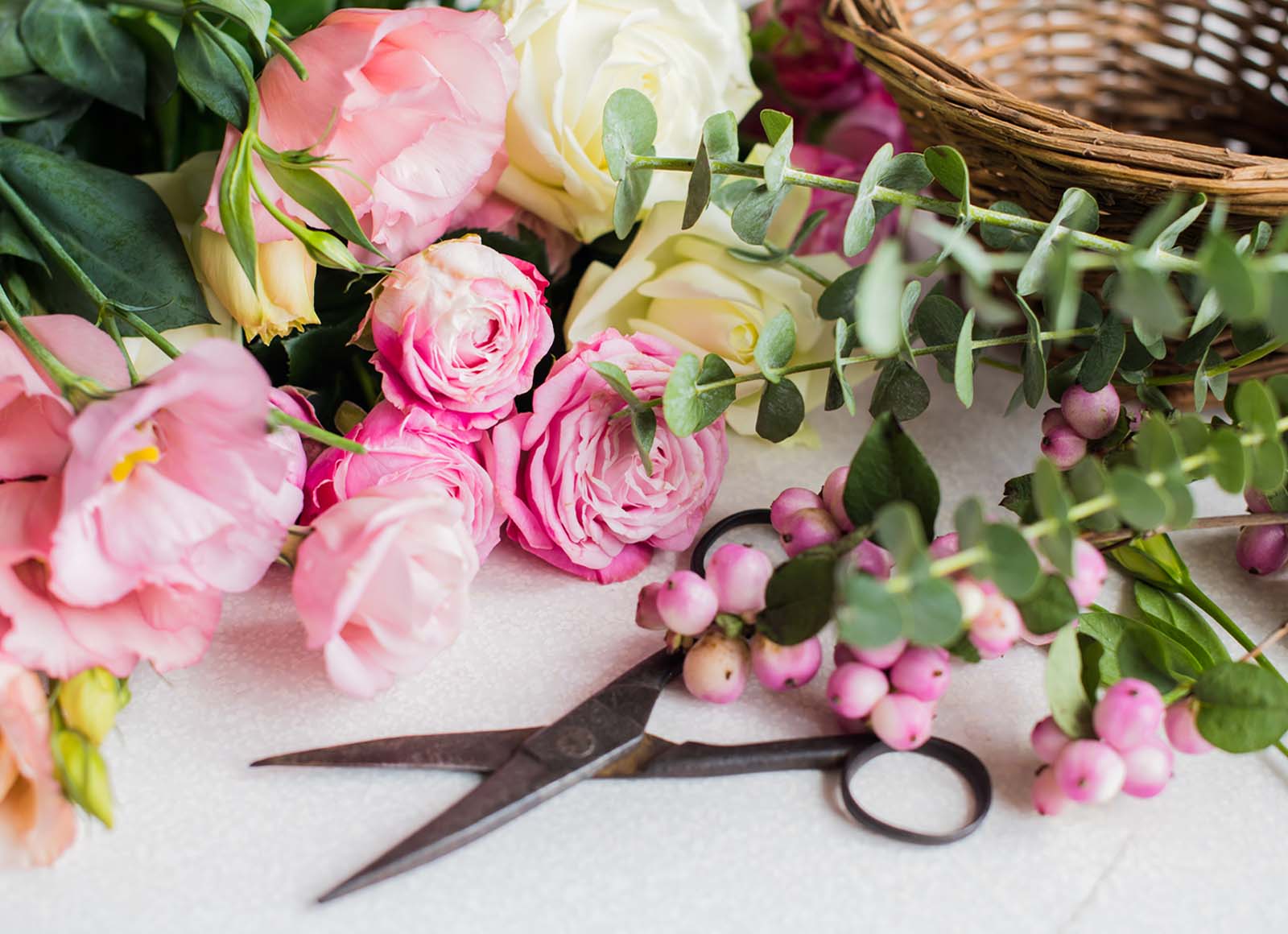 A close-up view of a craft surface shows pink and white flowers, greenery with pink berries, and open scissors, all arranged near a wicker basket on a white background.