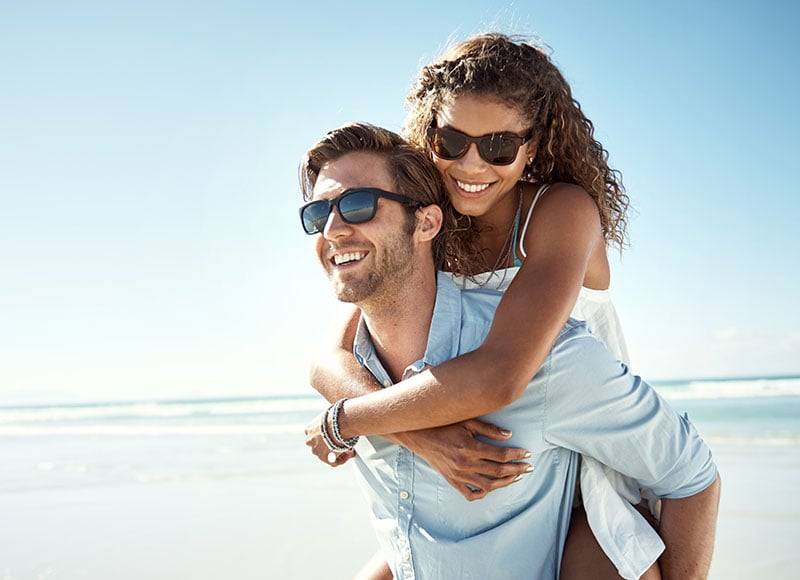 Husband gives wife a piggyback ride at the beach on a sunny day.