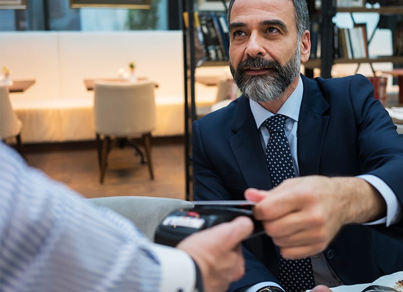 A man in a suit pays with a credit card at a restaurant, as a server's hand holds out a card reader to process the payment.