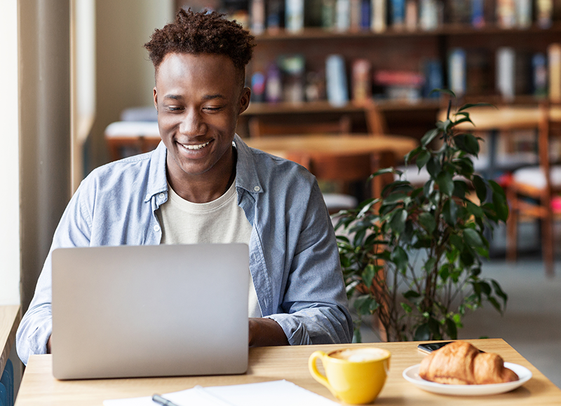 A young African-American man is sitting at a table in a coffee shop, smiling and looking at his laptop. He has a cup of coffee and a croissant on the table.