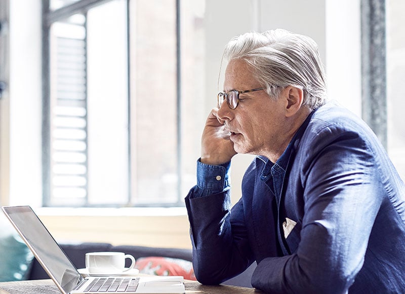 An older man sits at his desk and talks on the phone while reading something off of his laptop screen.