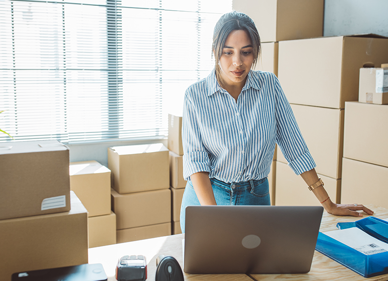 A young woman stands in a room full of boxes. She is wearing a blue and white striped shirt and jeans. She is looking at a laptop on a table.