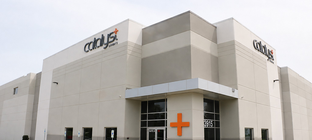 Exterior view of the Catalyst Exhibits office building featuring a modern beige and gray facade, large windows, the Catalyst logo on two corners of the building, and an orange plus sign displayed near the main entrance. A sidewalk, parking area, and landscaped lawn surround the building.