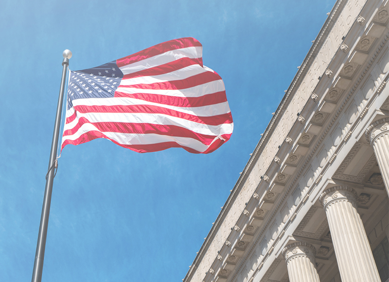 An American flag is waving in the wind against a blue sky. The flag is flying from a flagpole in front of a building with white columns.