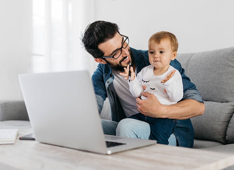 A dad sits on the couch with his baby while working on his laptop.