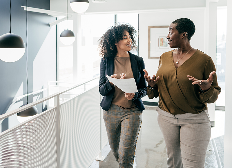 Two women are walking and talking in an office building. One woman is wearing a brown jacket and the other woman is wearing a black suit.
